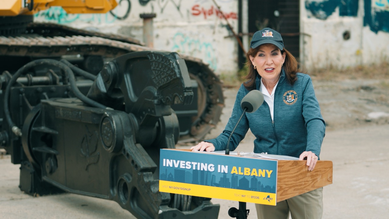 Woman in cap speaks at podium reading Investing in Albany, with tank in background