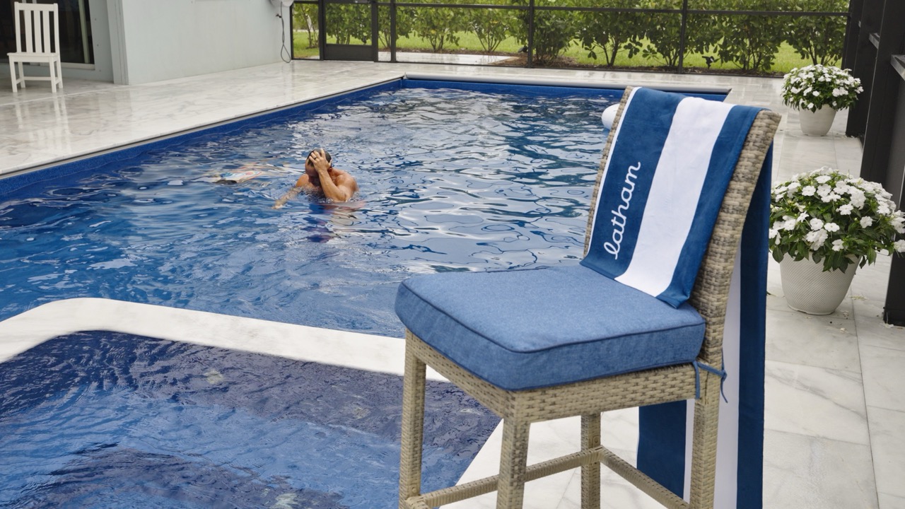 Man swimming in a blue pool beside a blue towel that reads Aquam, in a patio setting
