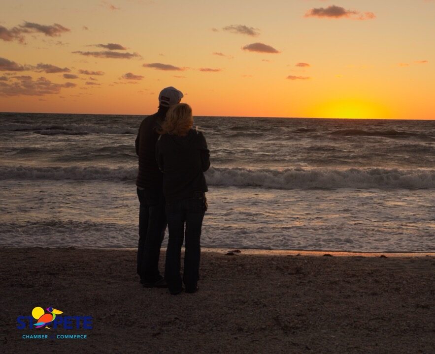 Couple in dark coats standing on beach at sunset with waves and clouds, logo text St. Pete Chamber Commerce